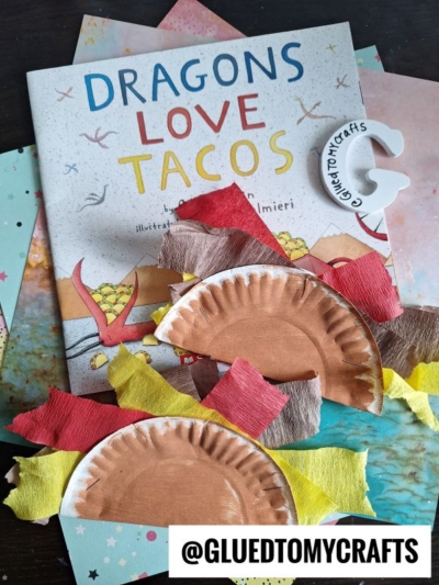 A stack of colorful paper, a Dragons Love Tacos book, dragon crafts made from painted paper plates and tissue paper, a Paper Plate Taco, and a @GLUETOMYCRAFTS tag are arranged on a dark surface.