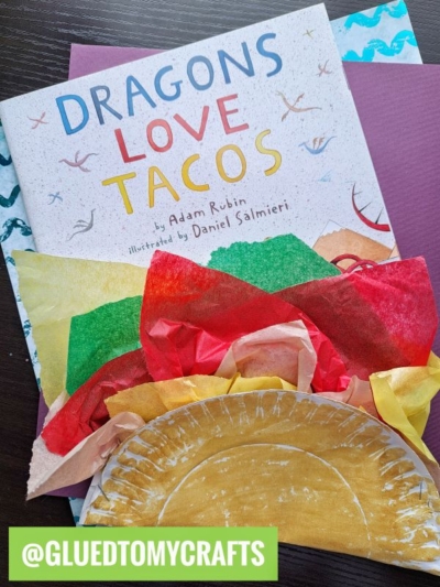 A copy of Dragons Love Tacos by Adam Rubin lies on a table, with a colorful paper plate taco craft made from tissue paper and a painted plate in front. A green @GLUEDTOMYCRAFTS tag is at the bottom.