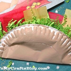 A paper plate taco painted brown and folded to resemble a taco, filled with crinkled green paper and pieces of red and yellow tissue for lettuce, tomatoes, and cheese. The crafty background features a website watermark.
