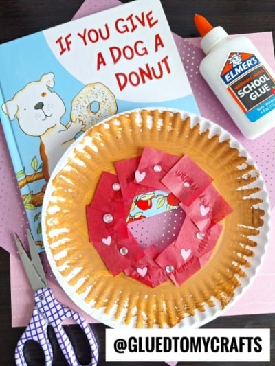 A children's book titled If You Give a Dog a Donut, Elmer's glue, scissors, and a Paper Plate Donut kid craft made with tissue paper and heart stickers are arranged on pink polka-dot paper.