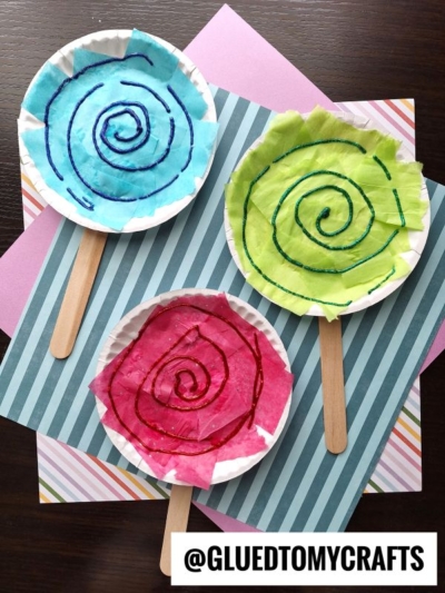 Three paper plate lollipop decorations with colorful tissue paper (blue, green, and pink) and glitter swirls sit on popsicle sticks, displayed against striped and pastel backgrounds. Fun and sweet paper plate lollipops for any craft day!.