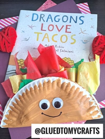 A children’s book titled “Dragons Love Tacos” is placed behind a paper plate taco craft decorated with googly eyes, a smile, and colorful tissue paper toppings. Yarn balls and red paper add to the fun taco-themed scene.