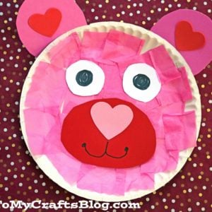 A pink bear face made from a paper plate, pink tissue paper, and paper cutouts for eyes, ears, and a heart-shaped nose looks adorable on a red polka dot background