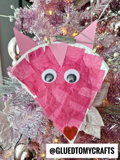 A pink cat face ornament, inspired by a Paper Plate Valentine Fox, is made from tissue paper, googly eyes, and pink paper ears and hangs on a light pink Christmas tree. A small red glitter heart decorates the bottom. The text @GLUEDTOMYCRAFTS appears below.
