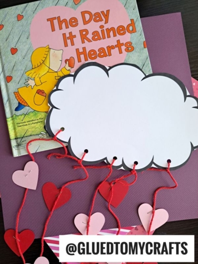 A children’s book titled The Day It Rained Hearts sits next to a tissue paper cloud craft, with red and pink paper hearts hanging from red string. @GLUEDTOMYCRAFTS is written at the bottom.