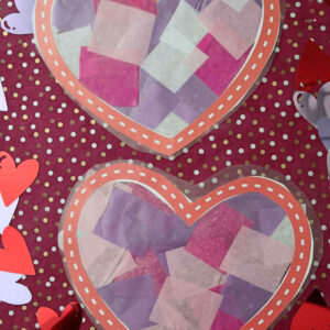 Two paper hearts filled with pink, purple, and white tissue squares create a stained glass hearts effect on a purple polka dot background, surrounded by cut-out hearts. Text reads “Stained Glass Hearts” and “GluedToMyCraftsBlog.com.”.