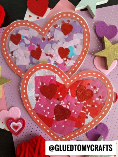 Two heart-shaped Stained Glass Hearts Valentine crafts, made with red borders, tissue paper pieces, and glittery red heart stickers, are displayed on a polka dot background surrounded by felt and foam heart and star decorations.