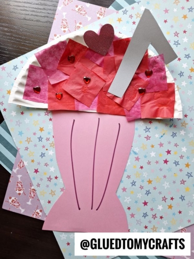 A colorful Paper Plate Milkshake craft with a pink cup, red and pink tissue paper whipped cream, heart-shaped gems, a red heart, and a silver straw, placed on patterned paper backgrounds.