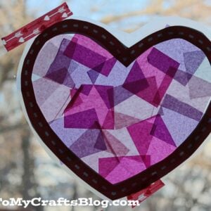 A heart-shaped suncatcher inspired by Stained Glass Hearts, made of overlapping pink and purple tissue paper, is taped to a window with decorative tape. GluedToMyCraftsBlog.com is visible at the bottom. Trees can be seen outside.