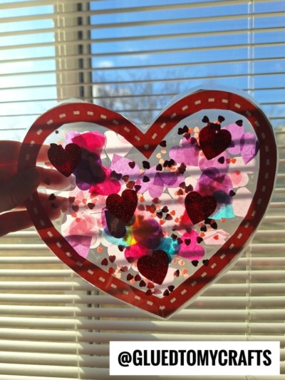 A hand holds a heart-shaped suncatcher, inspired by Stained Glass Hearts, with colorful tissue paper and red glitter hearts in front of window blinds. Sunlight shines through as @GLUEDTOMYCRAFTS appears at the bottom.