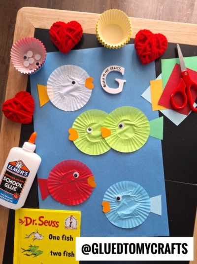 A flat lay of a Dr. Seuss-themed craft featuring fish made from colorful cupcake liners on a blue background. Each fish has a googly eye, an orange heart-shaped mouth, and a rectangular tail. The scene includes supplies like Elmer's glue, scissors, googly eyes, and yarn hearts, with a copy of "One Fish Two Fish Red Fish Blue Fish" by Dr. Seuss. The watermark "@GLUEDTOMYCRAFTS" is visible.