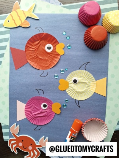 A colorful Cupcake Liner Rainbow Fish craft made with cupcake liners, paper cutouts, googly eyes, and sequins is displayed on blue paper with a glue stick. Decorative fish and crab cutouts add charm. @GLUEDTOMYCRAFTS is written at the bottom.