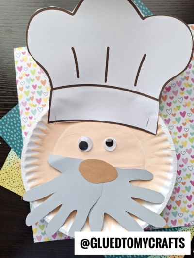 A paper plate baker craft featuring a chef’s face with a handprint beard, big chef hat, googly eyes, and brown oval nose, displayed on colorful patterned paper. @GLUEDTOMYCRAFTS is written at the bottom.