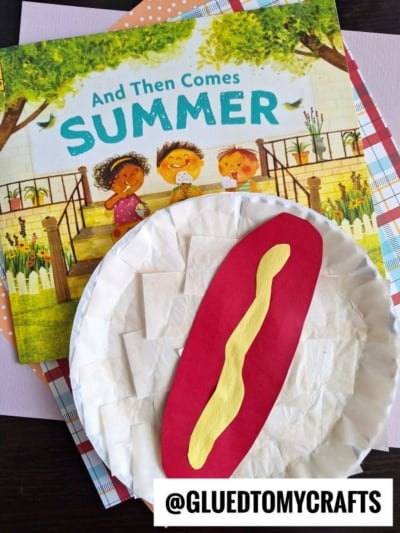 A colorful children’s book titled And Then Comes Summer is shown behind a creative Paper Plate Hot Dog craft with a red bun and yellow mustard. The username @GLUEDTOMYCRAFTS appears at the bottom.