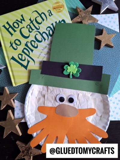 A children’s book titled How to Catch a Leprechaun sits next to a Paper Plate Leprechaun face craft, complete with a green hat, orange beard, and googly eyes. Gold stars and blue patterned paper decorate the background.