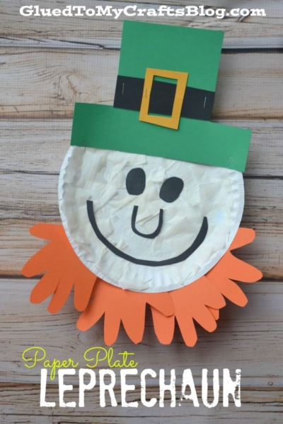 A cheerful Paper Plate Leprechaun craft with an orange paper beard and a green hat, featuring a smiling face, is displayed on a wooden background.