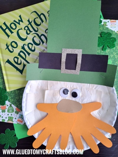 A St. Patrick’s Day craft featuring a Paper Plate Leprechaun face with orange handprint cutouts for a beard, googly eyes, and a paper hat, displayed on top of the book How to Catch a Leprechaun.