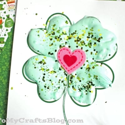A puffy paint shamrock of green with gold glitter and a pink felt heart in the center sits on white paper, surrounded by green patterned and festive papers.