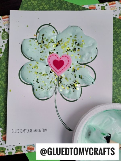 A Puffy Paint Shamrock outline filled with pale green puffy paint, topped with gold glitter and a pink felt heart in the center. The art is on printed paper with GLUEDTOMYCRAFTSBLOG.COM at the bottom.