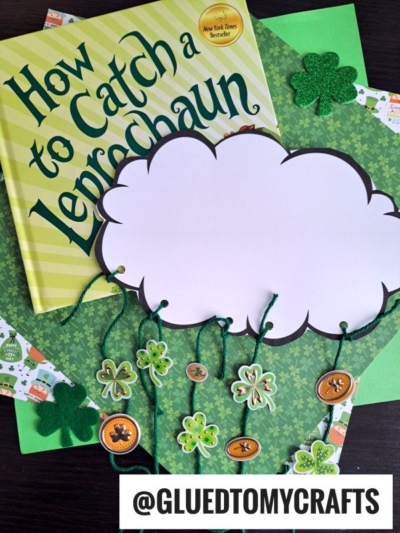 A copy of the book How to Catch a Leprechaun is surrounded by green craft paper, shamrock decorations, and a cloud-shaped cutout with strings and shamrock charms, creating a Raining Luck theme. The @GLUEDTOMYCRAFTS handle appears at the bottom.