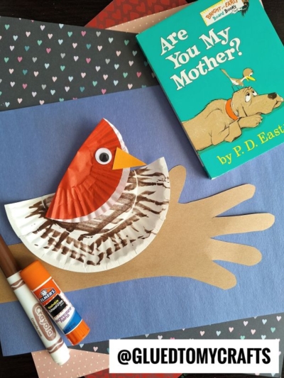A children’s craft project shows a bird made from a painted coffee filter and paper, placed on a cutout hand. Nearby are a glue stick, marker, and the book Are You My Mother? by P.D. Eastman—perfect for Dr. Seuss Kid Crafts inspiration.