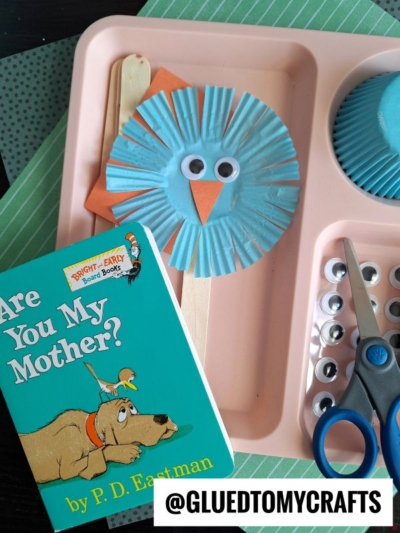 A children’s craft project inspired by Are You My Mother? shows a blue cupcake liner bird with googly eyes and a beak on a popsicle stick—perfect for fans of Dr. Seuss Crafts—beside supplies and the book cover on a pink tray.