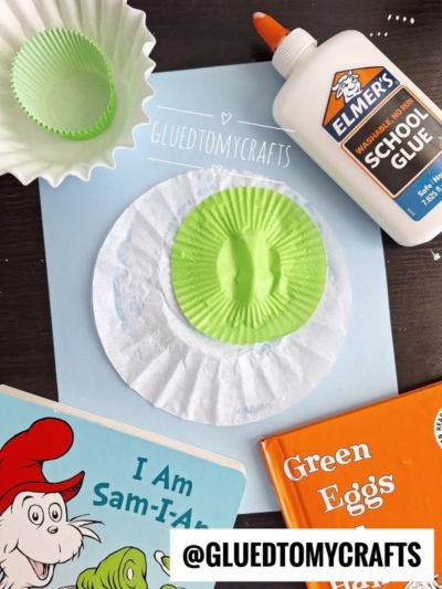 A paper craft setup inspired by Green Eggs and Ham with a book, glue, cupcake liners, a coffee filter, and a blue sheet labeled “GLUEDTOMYCRAFTS.” Perfect for Dr Seuss Kid Crafts, the green cupcake liner resembles green eggs.
