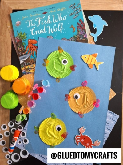 A children’s craft scene with a blue sheet featuring Cupcake Liner Rainbow Fish made from colorful cupcake liners, googly eyes, and paper fins. Nearby are googly eyes, glue, markers, sea animal cutouts, and The Fish Who Cried Wolf book.