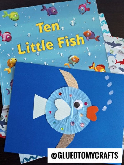 A children’s book titled Ten Little Fish is shown behind a blue card featuring a Cupcake Liner Rainbow Fish craft, decorated with sequins and paper cutouts. The handle @GLUEDTOMYCRAFTS is visible at the bottom.