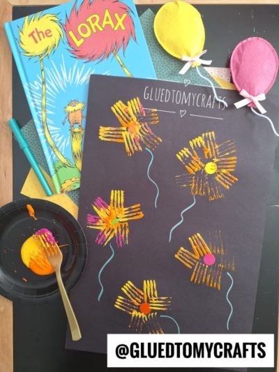 A creative Dr. Seuss Kid Craft inspired by “The Lorax,” featuring yellow and pink Truffula trees made with paint and fork prints on black paper. Nearby are a copy of “The Lorax” book, paint, and a fork for crafting fun.