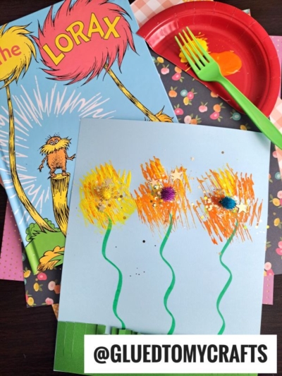 A children’s craft inspired by “The Lorax” features a book, a blue paper with colorful pom-pom flowers and green stems, and a red plate with yellow and orange paint—perfect for Dr. Seuss Crafts fans. Username @GLUEDTOMYCRAFTS is at the bottom.