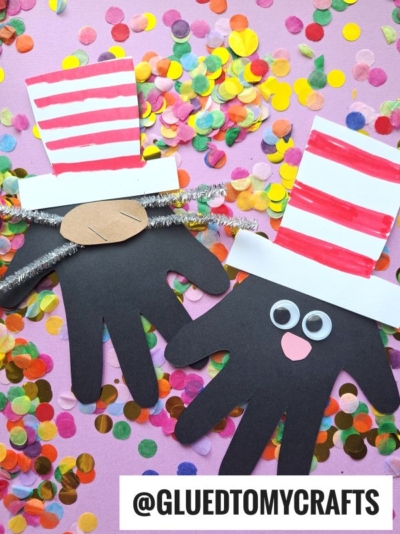 Two handprint crafts resembling the Cat in the Hat, with red and white striped hats, googly eyes, and pipe cleaner whiskers, placed on a pink background with colorful confetti—perfect for Dr Seuss Kid Crafts fun! @GLUEDTOMYCRAFTS text at the bottom.
