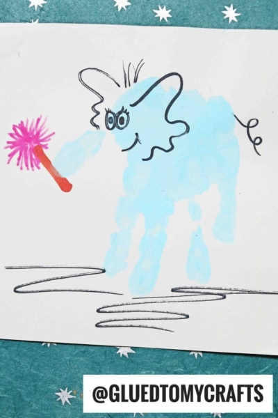 A blue elephant painted from a handprint holds a pink duster, with cartoon eyes and ears drawn on, perfect for Dr. Seuss Crafts. It stands on white paper with a teal star-patterned background. Text at the bottom reads @GLUEDTOMYCRAFTS.