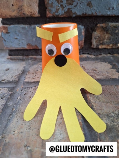 An orange paper cup Dr. Seuss Kid Craft, decorated with googly eyes, yellow paper eyebrows, and a yellow handprint beard to resemble a face, sits on a brick surface. @GLUEDTOMYCRAFTS text is at the bottom.