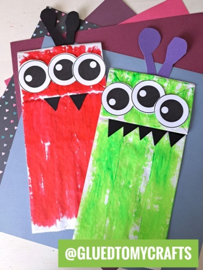 Two colorful Paper Bag Monster creations, one red and one green, each with three large googly eyes, black teeth, and two antennae, are displayed on stacked sheets of colored paper. A @GLUEDTOMYCRAFTS label is at the bottom.