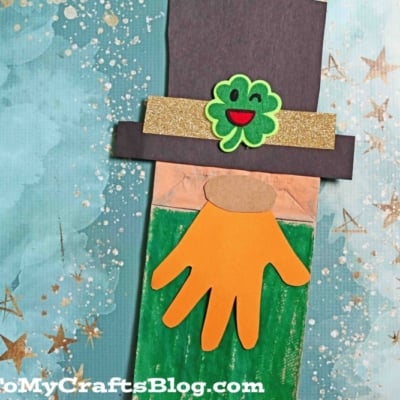 A paper bag leprechaun made of paper, featuring a black hat with a gold band and shamrock, an orange hand-shaped beard, and a green body, set on a blue background with gold stars.