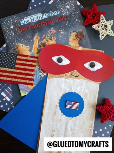 A children’s book titled The Night Before the Fourth of July, a Paper Bag Superhero puppet with a red mask and American flag sticker, red and white star decorations, and an Instagram handle @GLUEDTOMYCRAFTS.