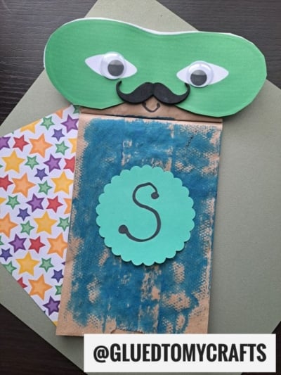 A Paper Bag Superhero puppet with a green mask, googly eyes, black mustache, blue painted mouth area, star-patterned cape, and a scalloped circle with an S. The handle @GLUEDTOMYCRAFTS appears at the bottom.