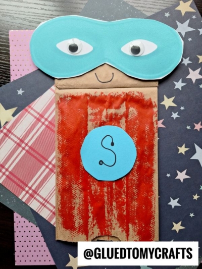 A Paper Bag Superhero puppet decorated with a blue mask, googly eyes, a smiling face, and a red painted body featuring a blue circle with an S. The paper superhero rests on star-patterned and plaid paper.