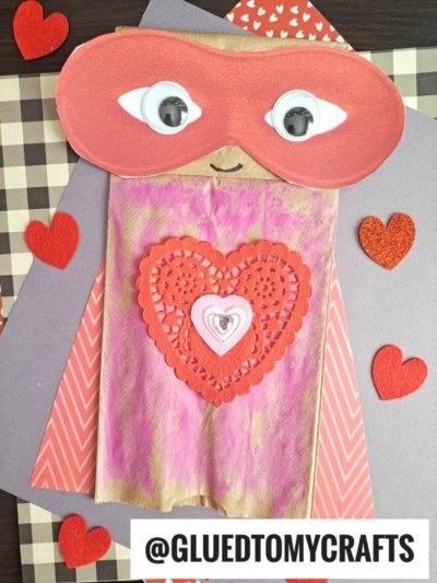 A Paper Bag Superhero puppet decorated with a red mask, googly eyes, a pink and red heart on its chest, and surrounded by red hearts. The handle @GLUEDTOMYCRAFTS appears at the bottom.