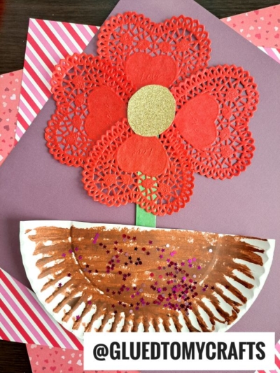 A red paper doily flower with a gold center and green stem is glued onto a pink background. The brown, glittery half-circle pot gives it a Paper Plate Love Plant vibe. GLUEDTOMYCRAFTS is written at the bottom.
