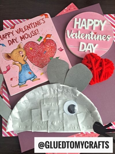 A handmade Paper Plate Mouse Valentine’s Day craft with a googly eye and paper ears sits on pink paper next to a Valentine card with a mouse, a red yarn heart, and a “Happy Valentine’s Day” sign. @gluedtomycrafts is at the bottom.