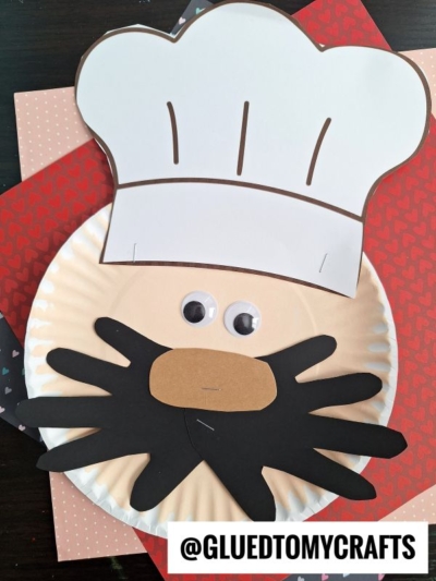 A Paper Plate Baker craft featuring a chef’s face with a white hat, googly eyes, and a black paper mustache and beard, placed on colored paper. The image includes the text “@GLUEDTOMYCRAFTS.”.