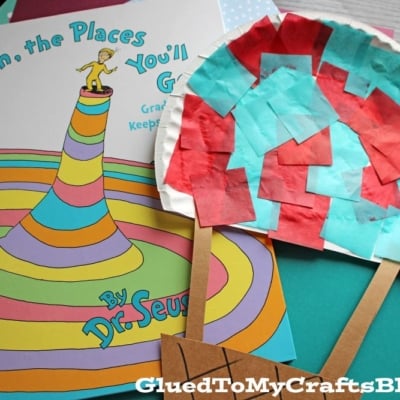 A copy of Seuss Oh The Places You'll Go! by Dr. Seuss sits next to a handmade craft resembling a popsicle, made of colored paper squares and sticks, on a dark surface—perfect for inspiring creativity like a Plate Hot Air Balloon activity.