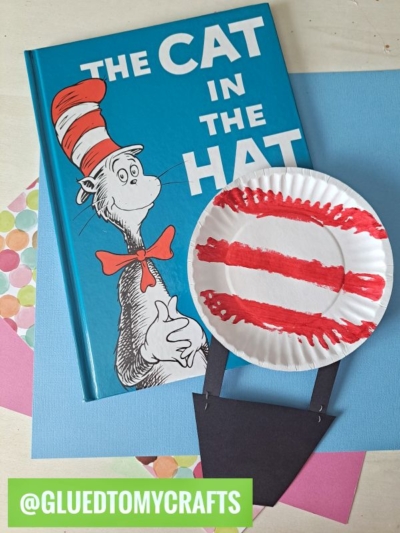 A copy of The Cat in the Hat book sits beside a paper plate craft resembling the cat’s hat, painted with red stripes and a black paper brim—perfect inspiration for fun Dr. Seuss Crafts. Colorful construction paper decorates the background.