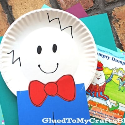 A cheerful Paper Plate Humpty Dumpty craft with a smiling face, zigzag hair, red bow tie, blue pants, tan legs, and red shoes sits on colorful paper, with a Humpty Dumpty book in the background.