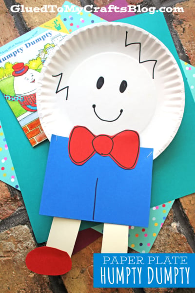 A cheerful Paper Plate Humpty Dumpty craft with a smiling face, zigzag hair, red bow tie, blue pants, tan legs, and red shoes sits on colorful paper, with a Humpty Dumpty book in the background.