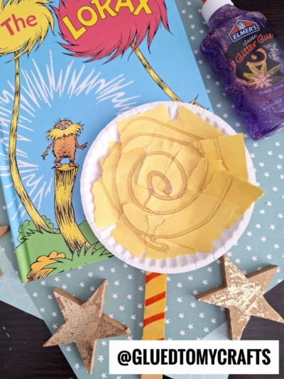 A Dr. Seuss Kid Crafts project inspired by The Lorax, this yellow paper plate flower uses torn paper, gold glitter glue, and a striped straw stem. Wooden stars and craft supplies are arranged nearby. The handle reads @GLUEDTOMYCRAFTS.