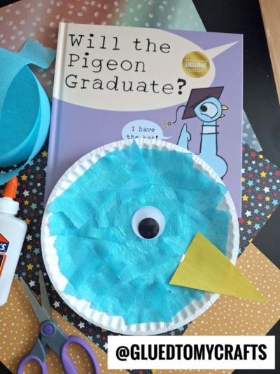 A children’s book titled Will the Pigeon Graduate? is shown with a colorful Paper Plate Pigeon craft made from tissue paper, a yellow beak, and a googly eye. Scissors, glue, and craft materials are nearby. @GLUEDTOMYCRAFTS is written below.