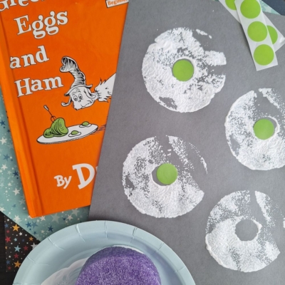 A copy of Green Eggs and Ham by Dr. Seuss sits beside a gray paper with white painted egg shapes and green circles—perfect for Dr Seuss Kid Crafts. Craft supplies and a purple sponge are nearby. @GLUETOMYCRAFTS is written at the bottom.